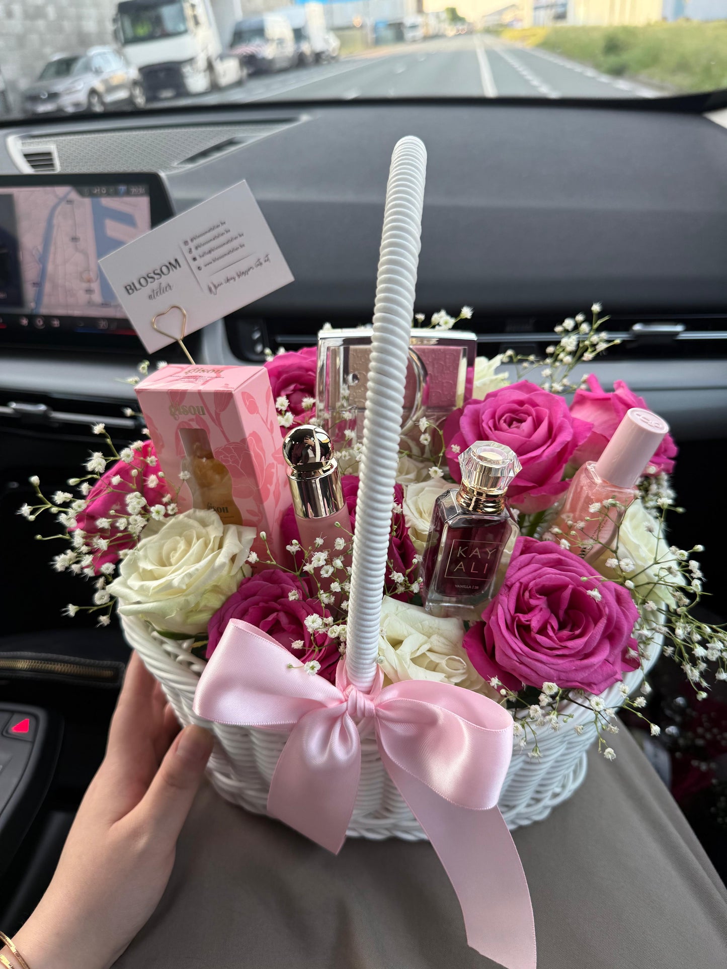 Floral arrangement with pink and white roses, a perfume bottle, and a small box inside a basket held by a person in a car.
