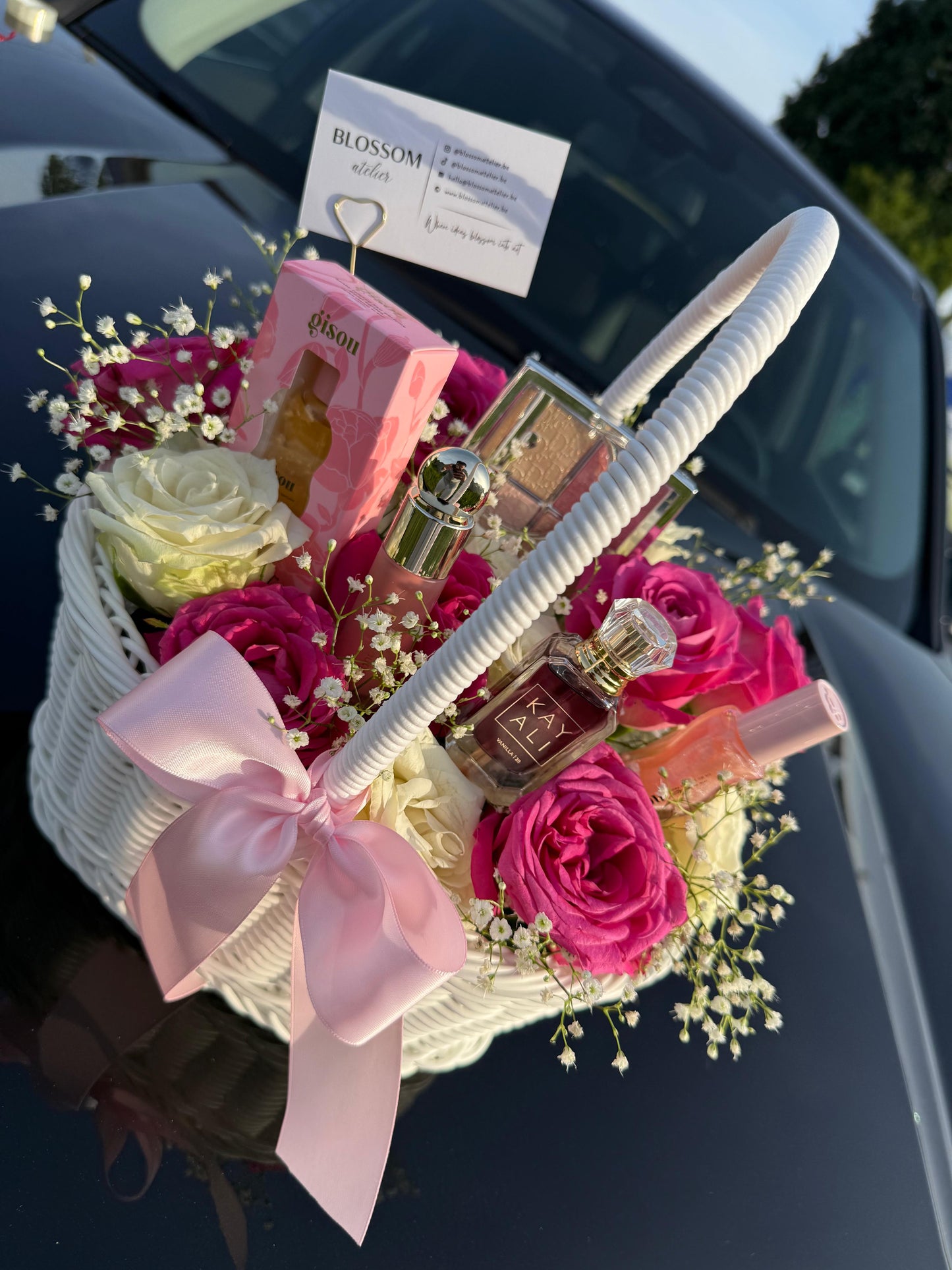 Basket of flowers and perfumes with a pink bow on a car hood