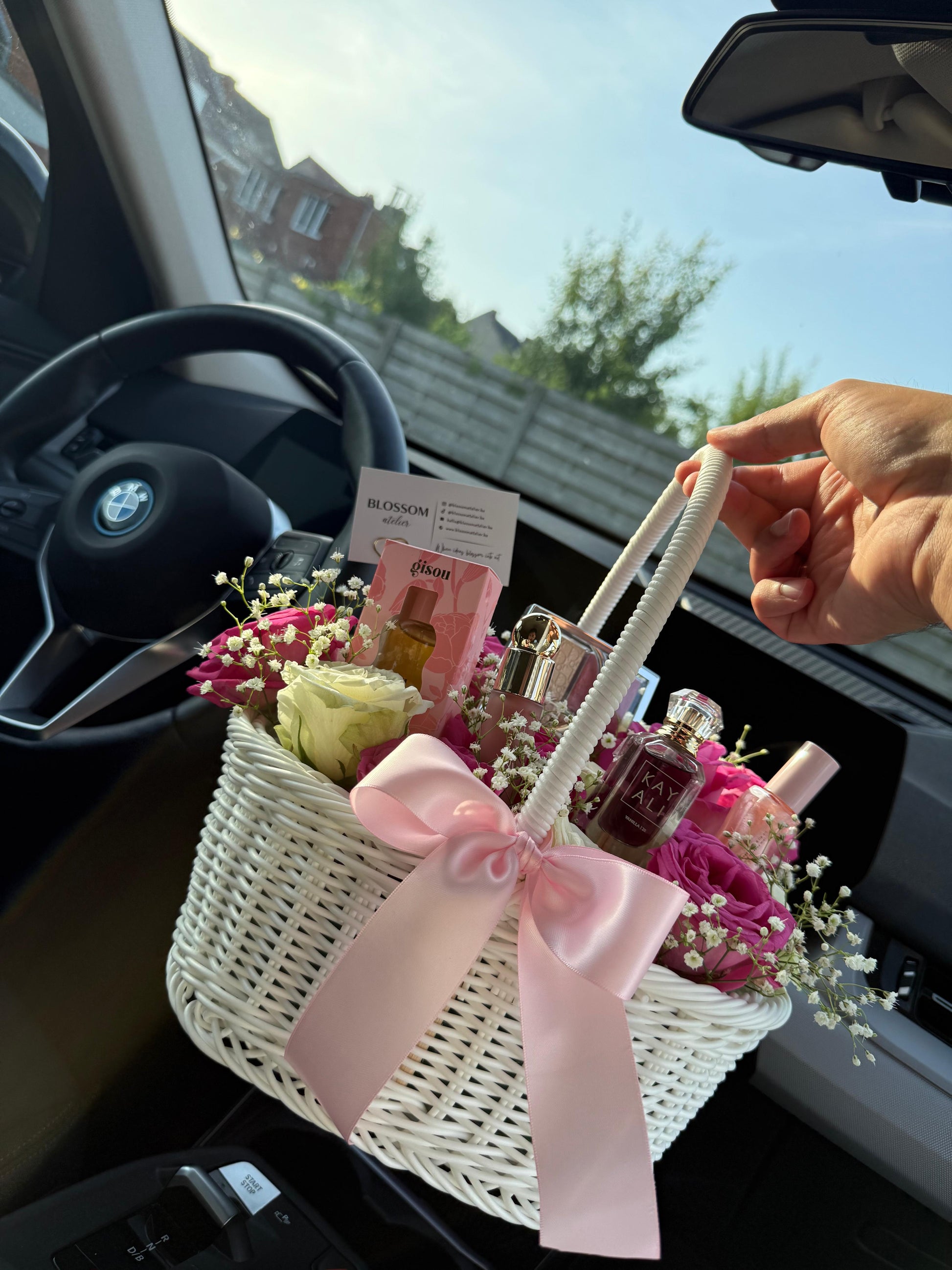White gift basket with flowers and perfumes held by a hand inside a car.